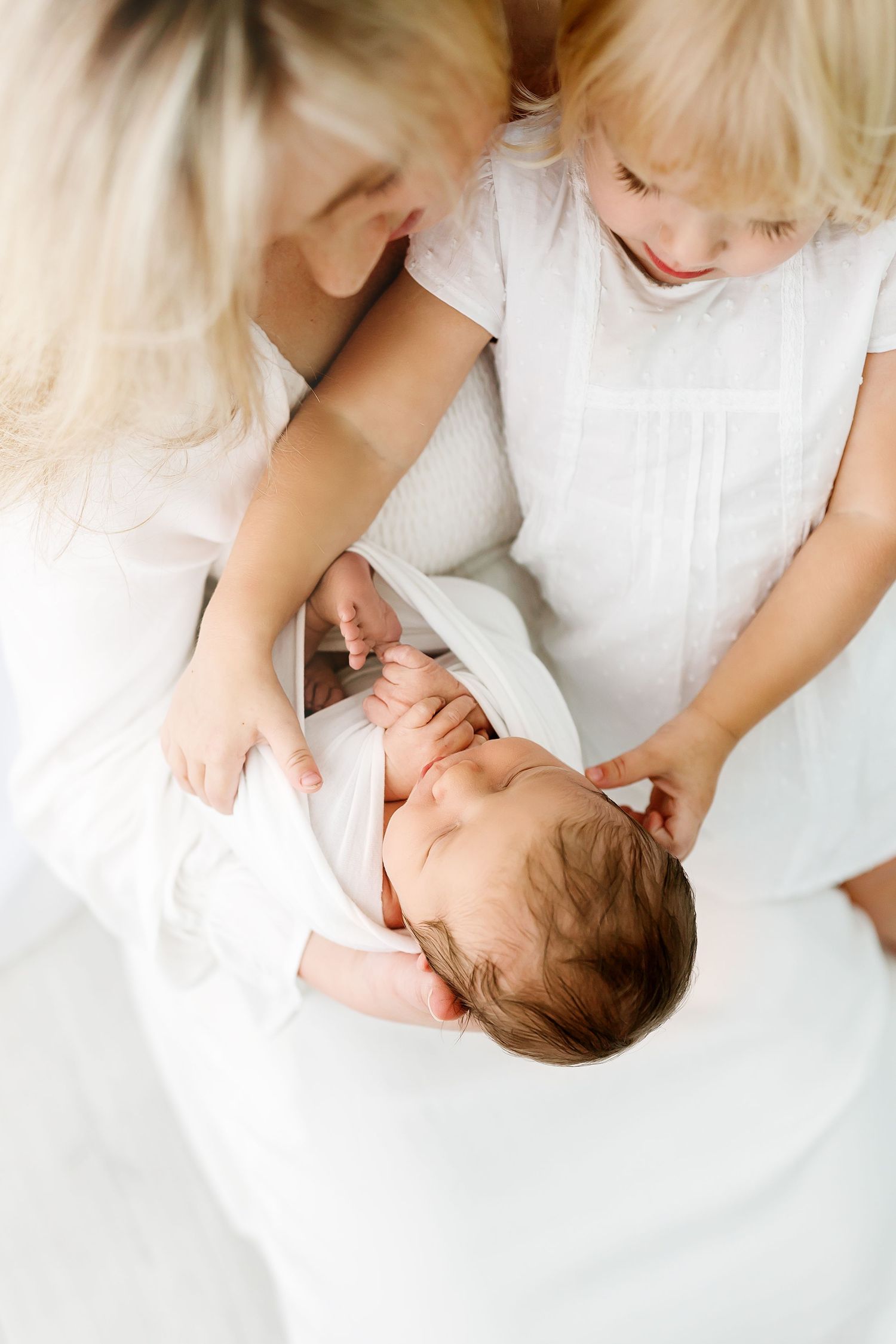 Parent in white holding and gazing lovingly at a newborn baby wrapped in blankets on white bedding.