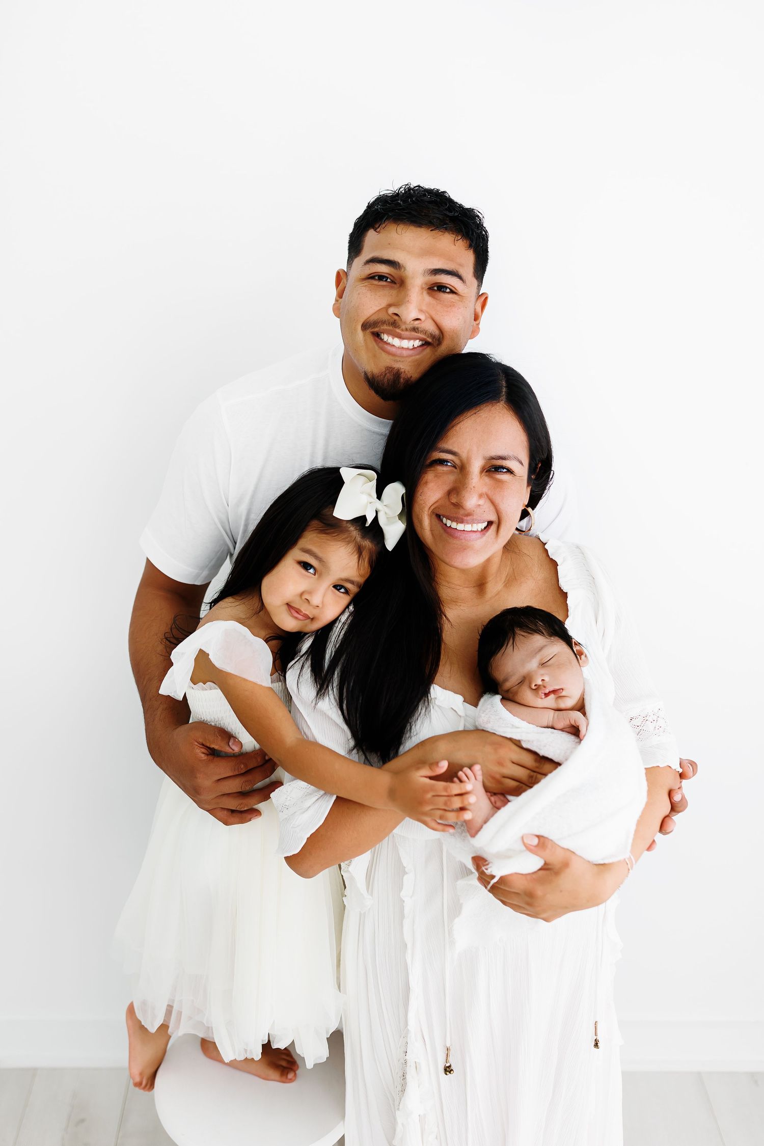 A family in matching white outfits laughs and embraces in a minimalist studio setting against a white background.