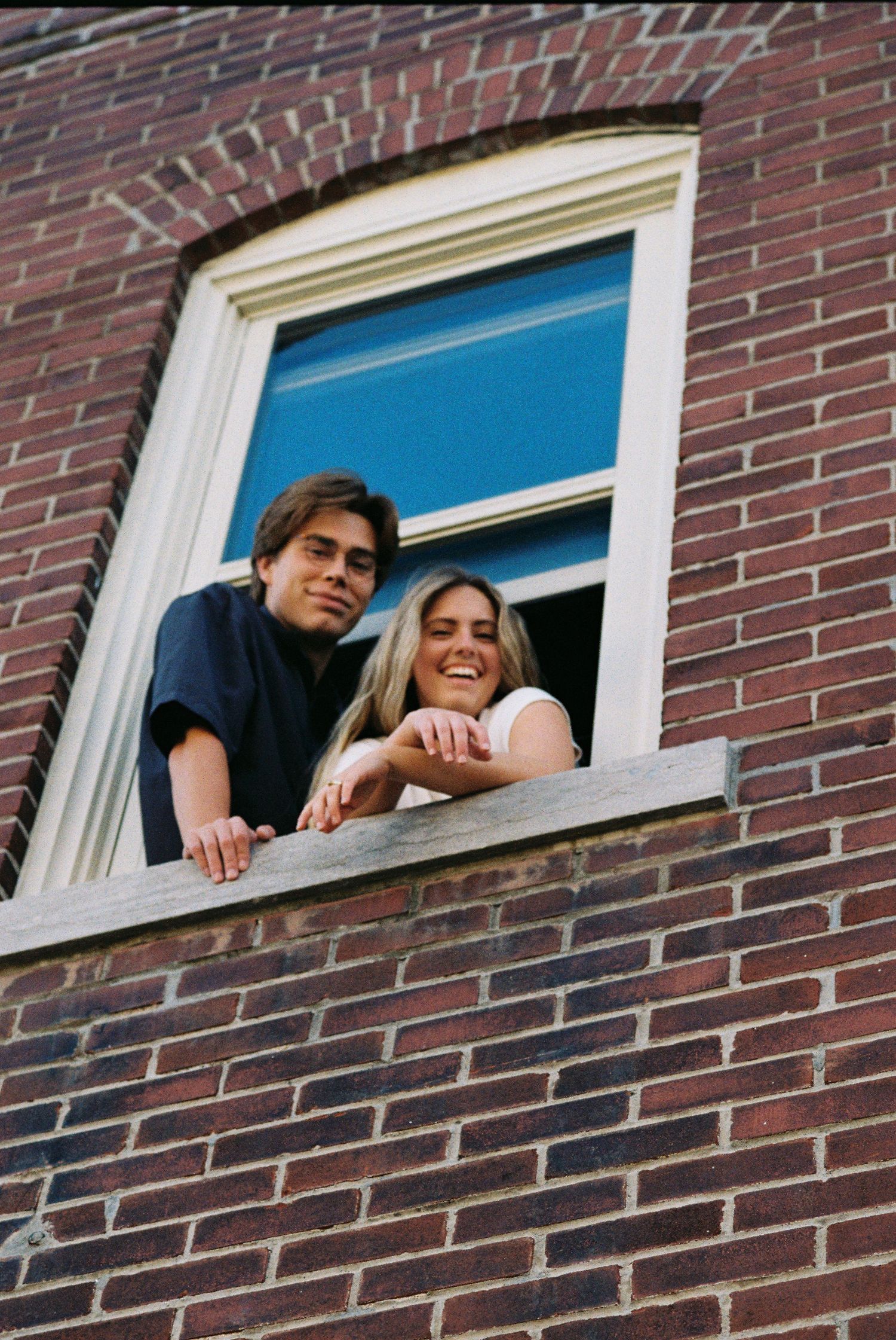 Two people lean out of a window frame in a brick building, sharing a casual moment together.