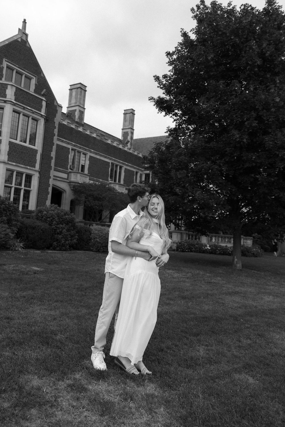 A couple embraces on a lawn in front of a stately Victorian mansion in black and white.