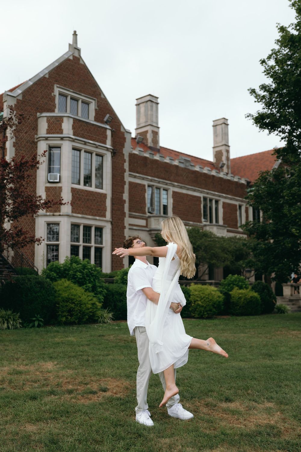 A couple shares a playful moment on the lawn of a grand brick mansion.