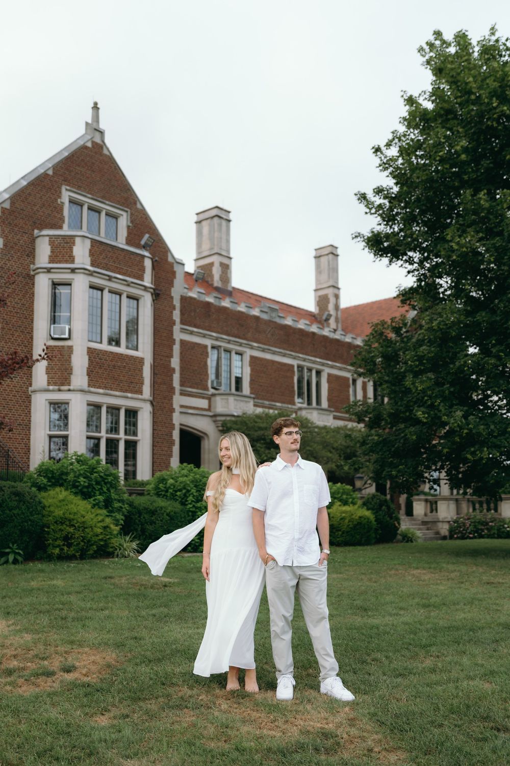A couple in coordinating white outfits poses on the lawn of a historic brick building.