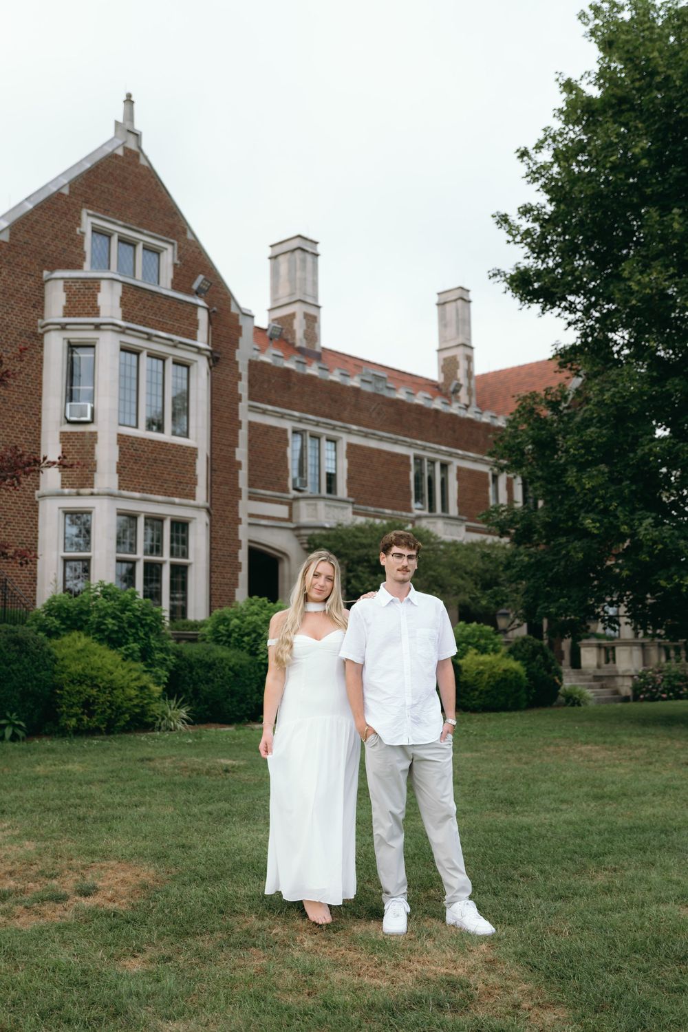 A couple in white stands together on the lawn of a historic brick mansion.