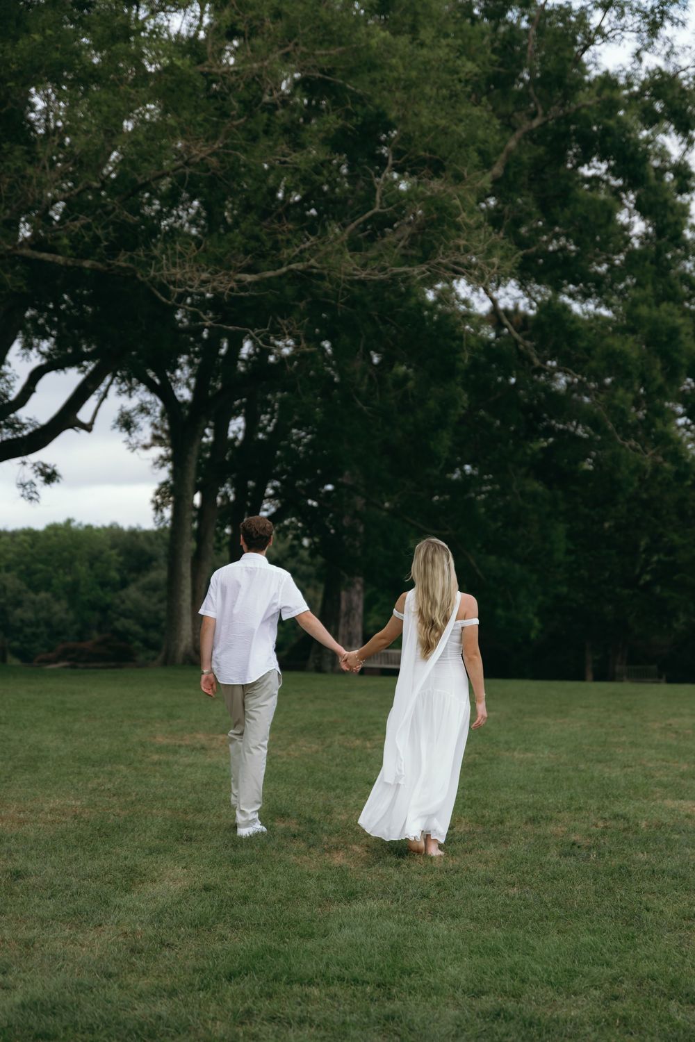 A couple walks hand in hand across a grassy field with trees in the background.