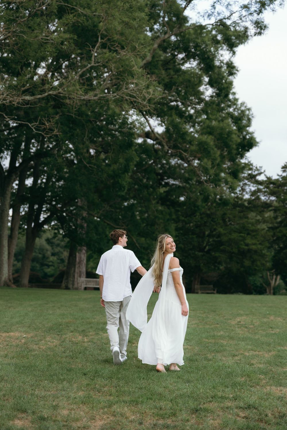 A couple in white attire walks together across a grassy field with trees in the background.