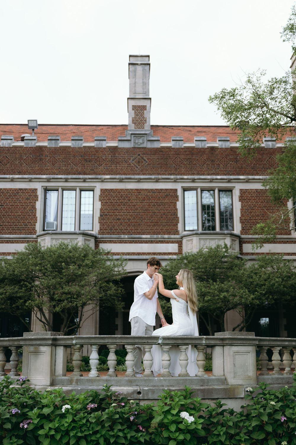 Two people in white stand near a brick mansion with ornate stone balustrades and landscaping.