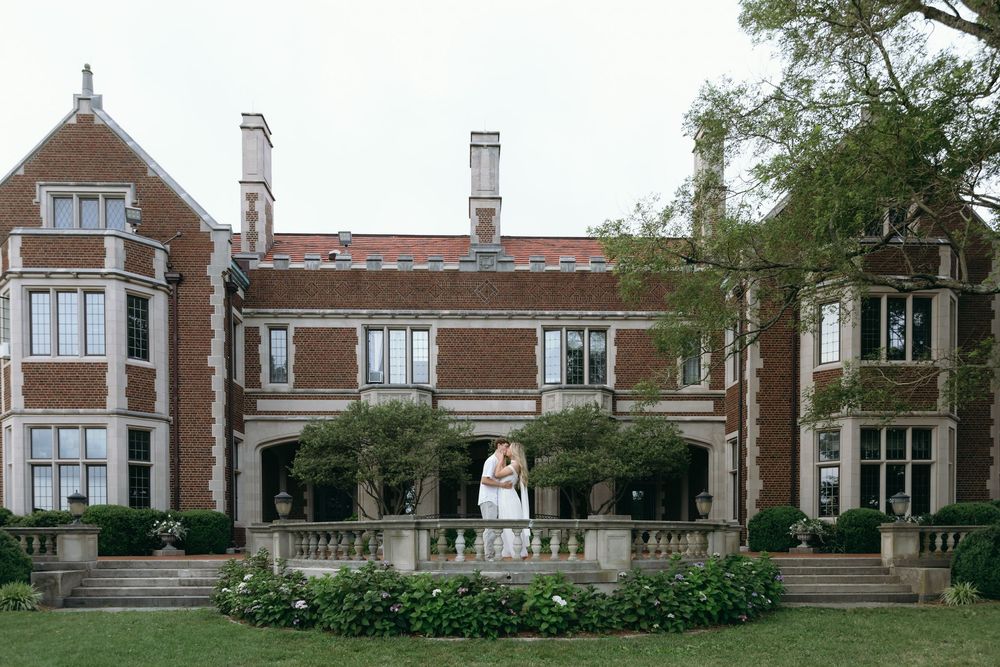 Wide panoramic view of a stately brick mansion with stone architectural details and manicured grounds.