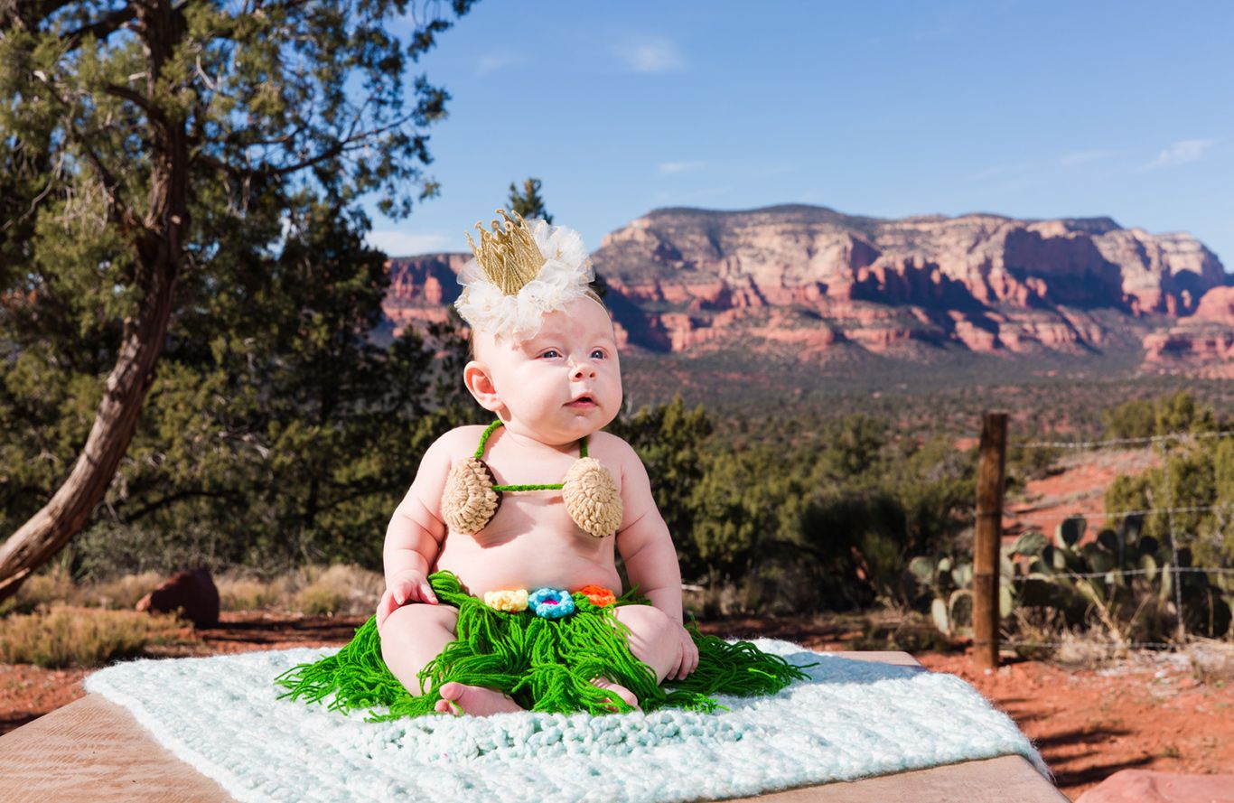 A baby sits on a blanket in the desert with red rock formations of Sedona visible in the background.