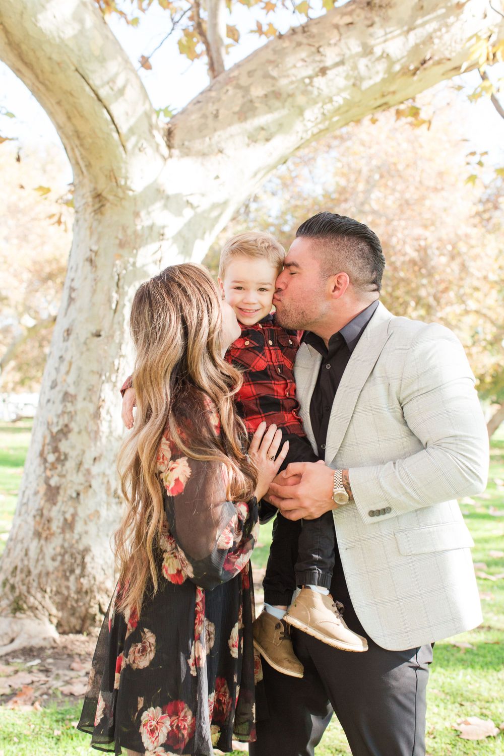 Family photo sequence under large tree branches with parents kissing child in floral dress.