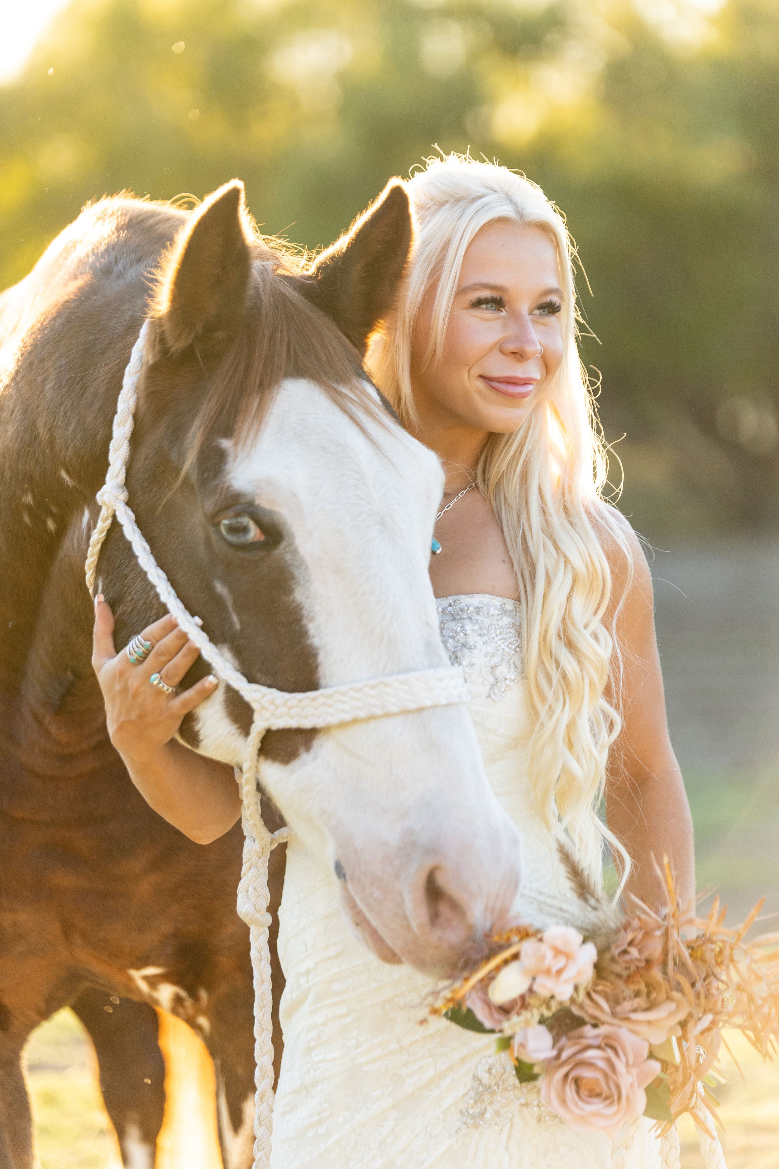blonde woman wearing a wedding dressing holding a bouquet standing next to a horse