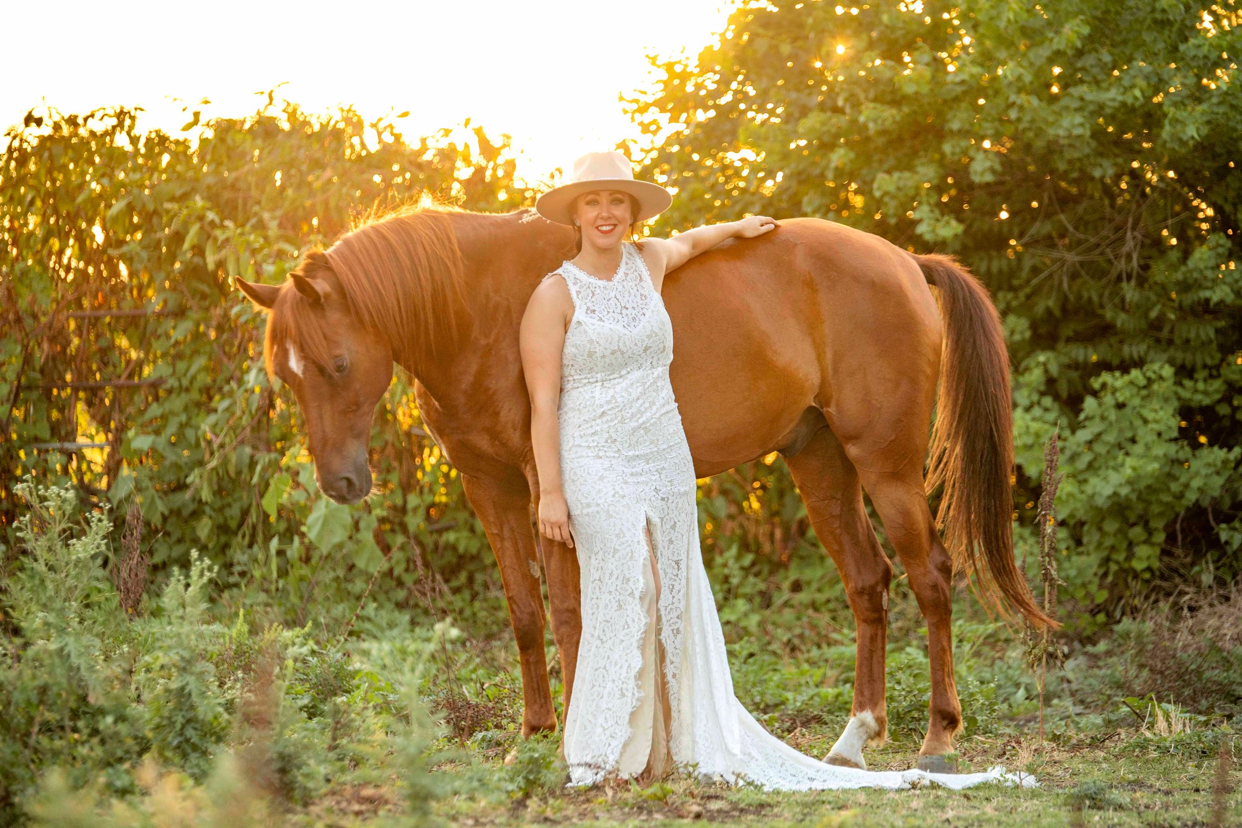 equestrian bridal portraits, woman standing next to her horse in a field at sunset wearing a white lace wedding dress.
