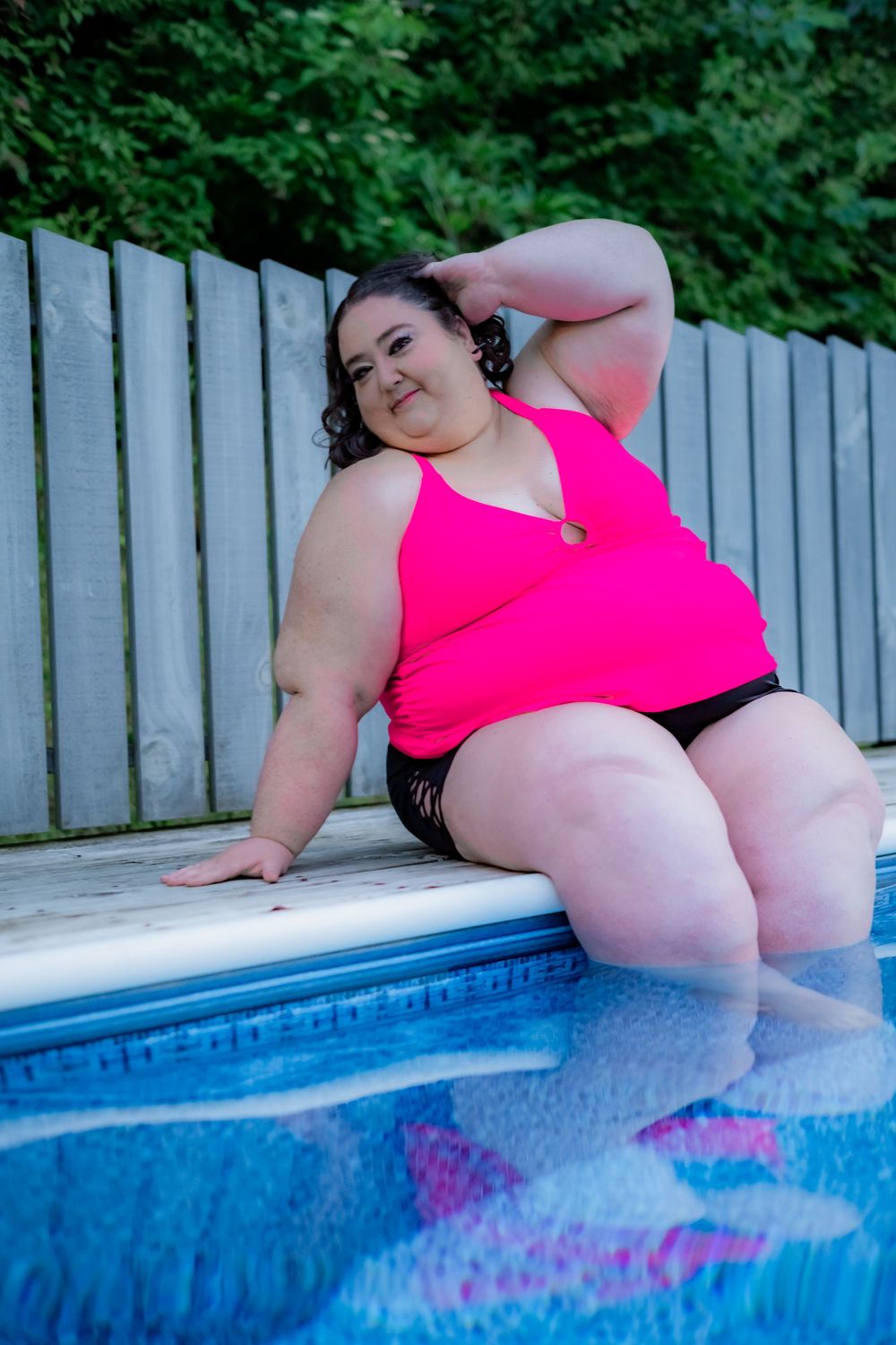 Plus size woman in pink tankini sitting poolside with feet in water, wooden fence and green trees in background. Pool/Poolside Boudoir.