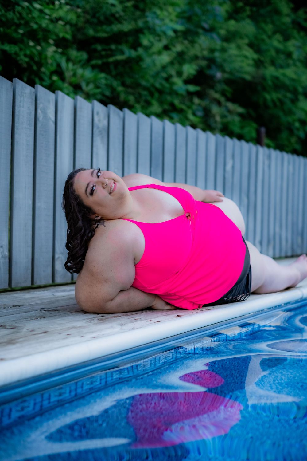 Plus-size woman in pink swimsuit posing poolside, smiling confidently by blue water with wooden fence backdrop. Pool/Poolside Boudoir.