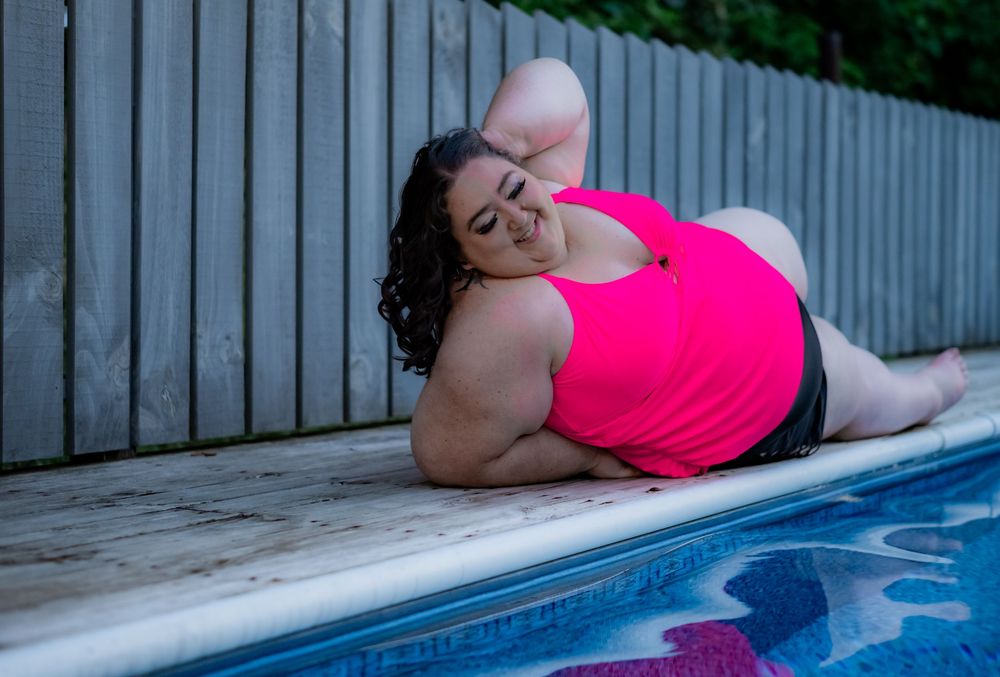 Plus-size woman in pink tank top posing by outdoor pool on wooden deck near wooden fence. Pool Boudoir. Poolside Boudoir.