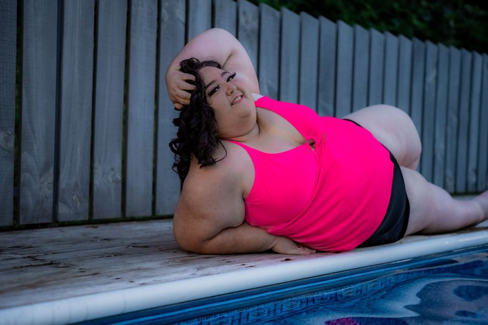 Plus-size woman in pink tank top relaxing by a swimming pool, posing confidently on the pool deck. Pool Boudoir. Poolside Boudoir.