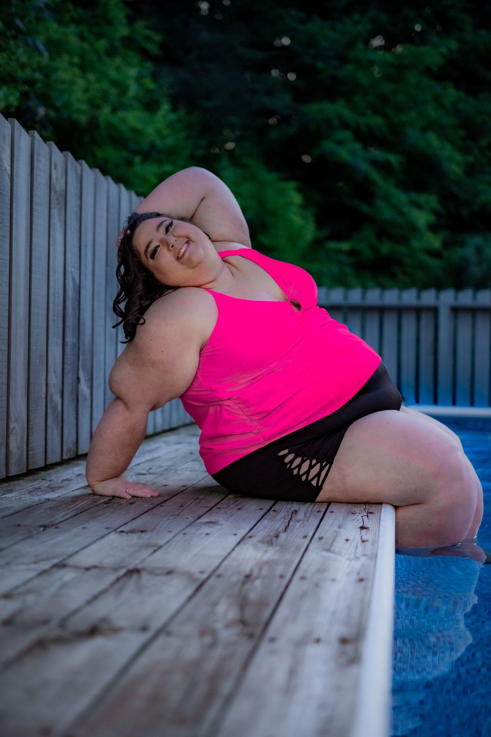 Plus-size woman in pink tank top and black shorts posing on wooden pool deck with green trees background. Pool Boudoir. Poolside Boudoir.