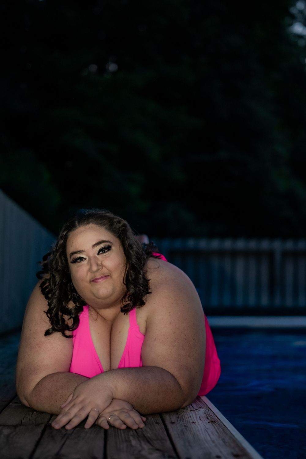 Plus-size woman in pink swimsuit posing poolside on wooden deck at dusk with dark trees in background. Pool Boudoir. Poolside Boudoir.