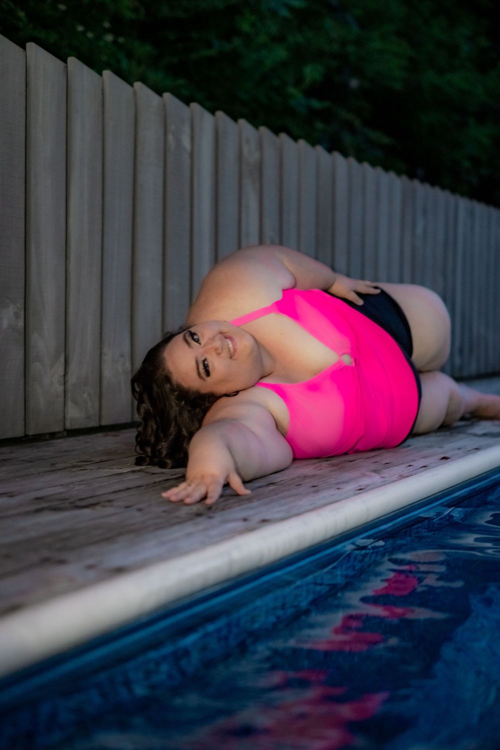 Plus-size woman in pink swimsuit posing by pool on wooden deck near fence. Pool Boudoir. Poolside Boudoir.