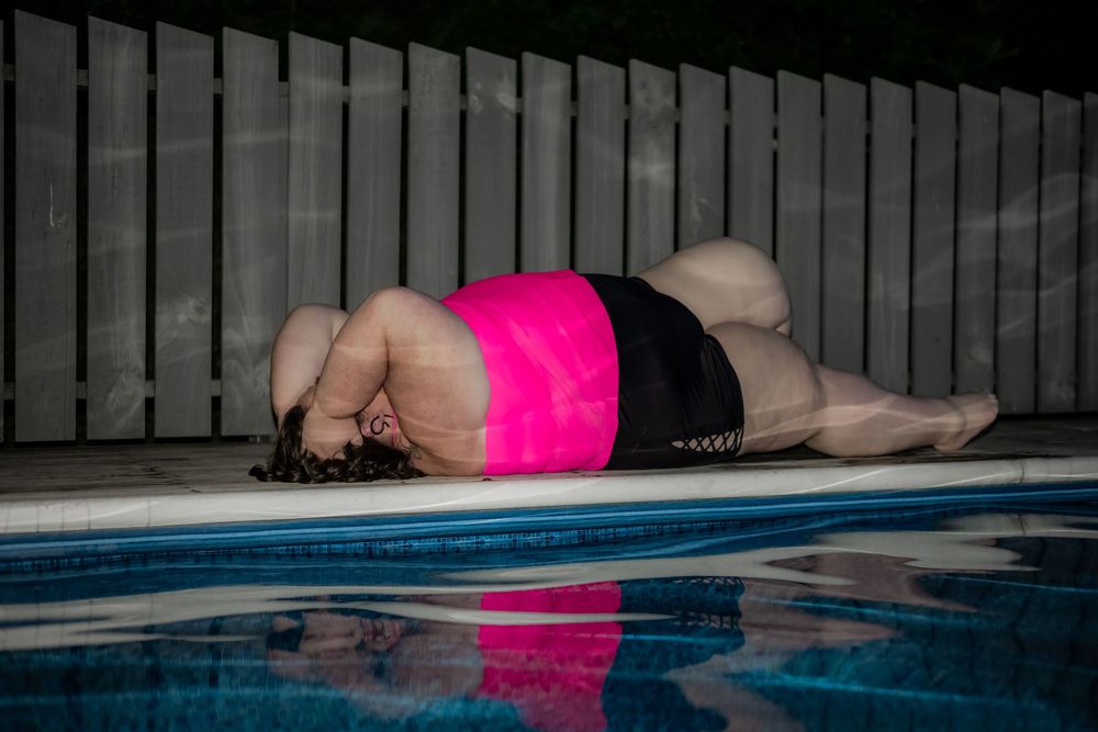 Person in pink top and black shorts lying by a pool at night near a wooden fence. Pool Boudoir. Poolside Boudoir.