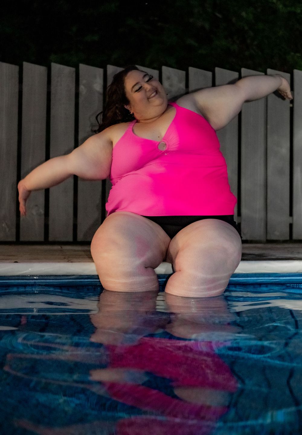 Plus size woman in pink & black swimsuit sitting at the edge of a swimming pool during blue hour at night. Pool Boudoir. Poolside Boudoir.