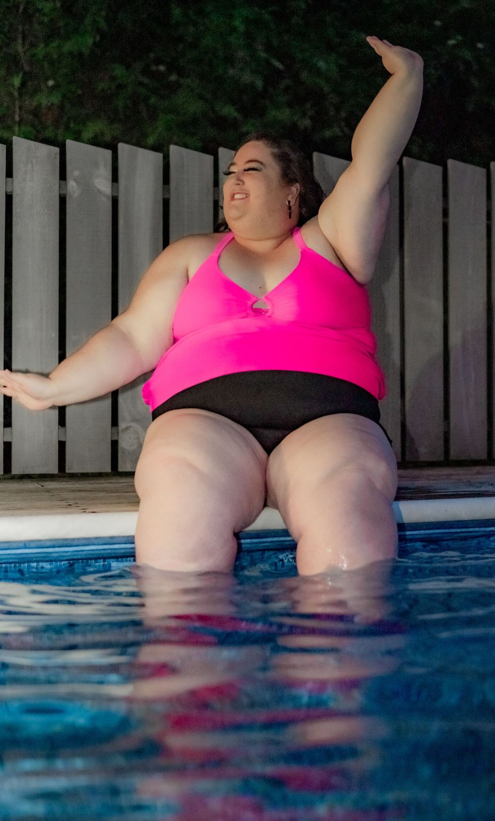 Plus size woman in pink & black swimsuit sitting at the edge of a swimming pool during blue hour at night. Pool Boudoir. Poolside Boudoir.
