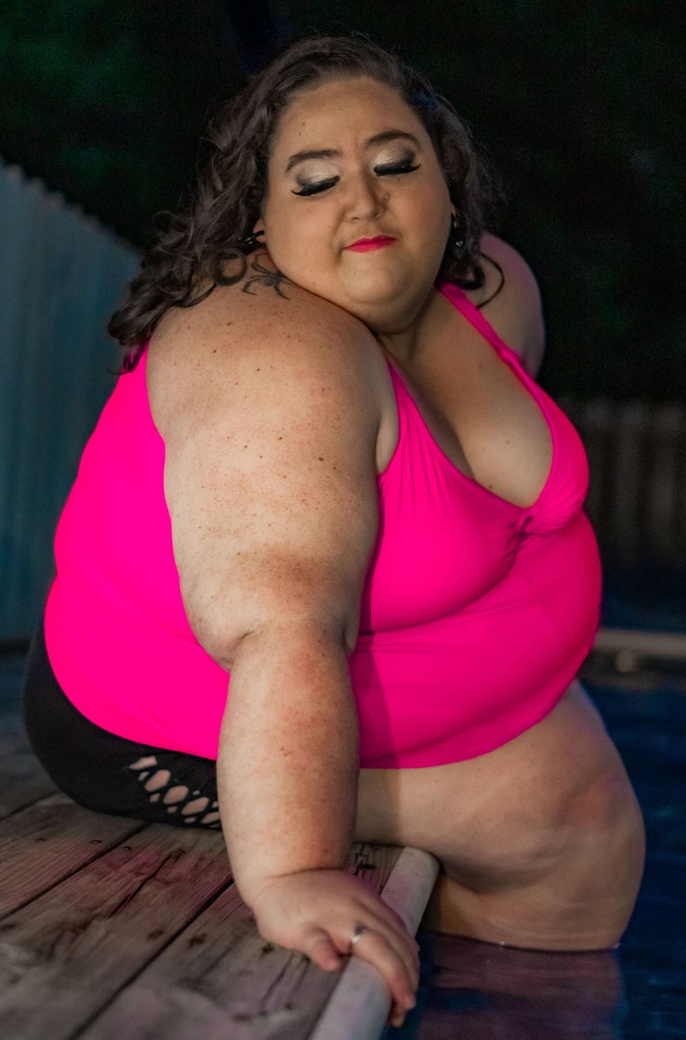 Plus-size woman in pink bikini top posing confidently by a pool on a wooden deck outdoors. Pool Boudoir. Poolside Boudoir.