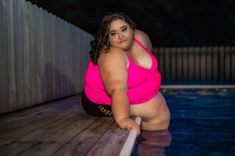 Plus-size woman in pink bikini top posing by a pool at night, leaning on the edge with confidence. Pool Boudoir. Poolside Boudoir.