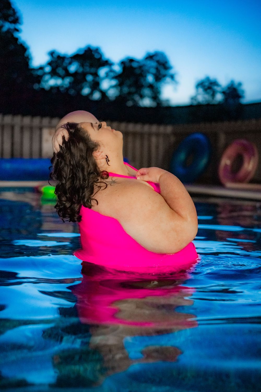 Woman in pink bikini relaxing in a pool at dusk, with colorful inflatable rings visible in the background. Pool Boudoir. Poolside Boudoir.