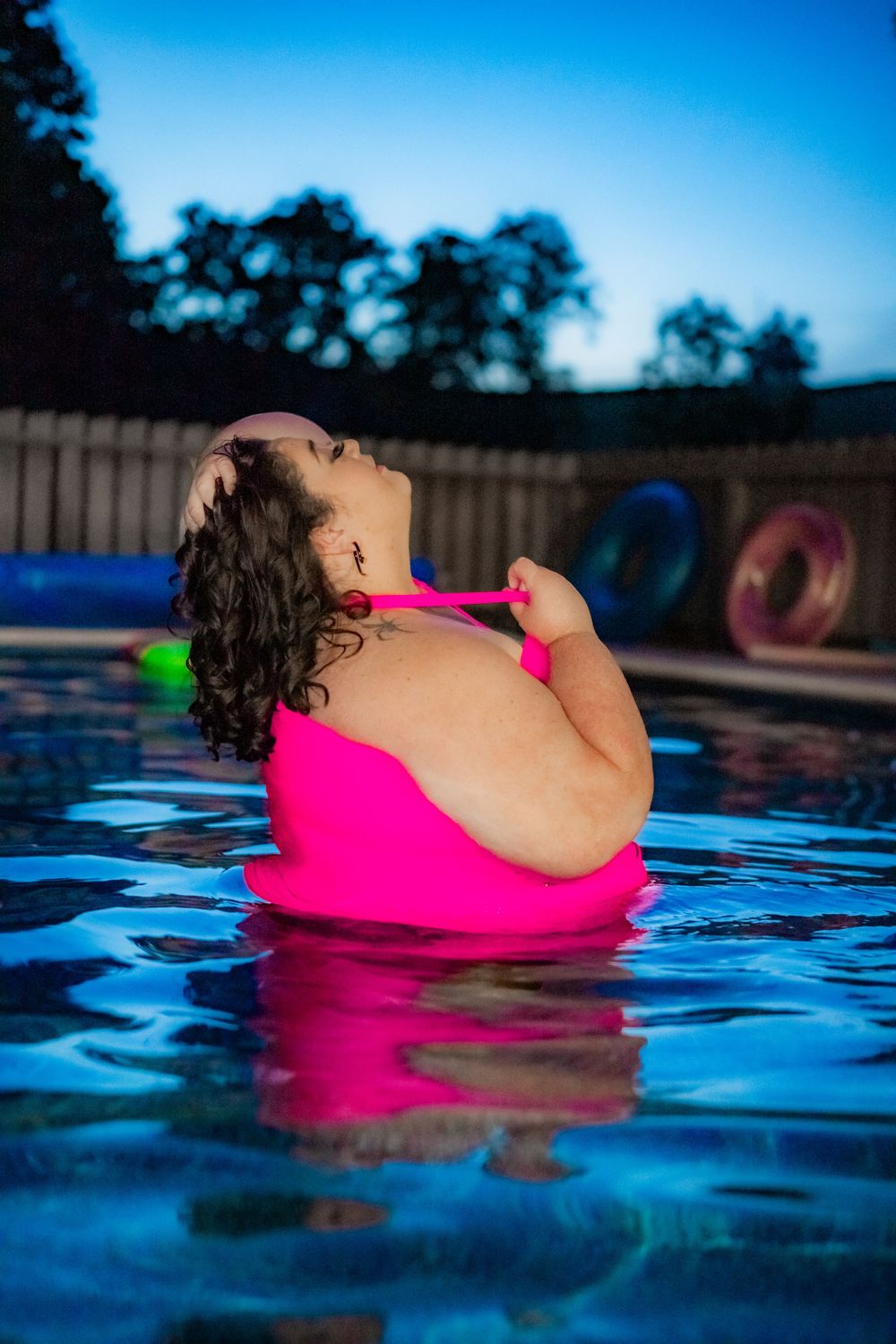 Woman in pink swimsuit relaxing in illuminated pool at dusk with colorful floats in background. Pool Boudoir. Poolside Boudoir.