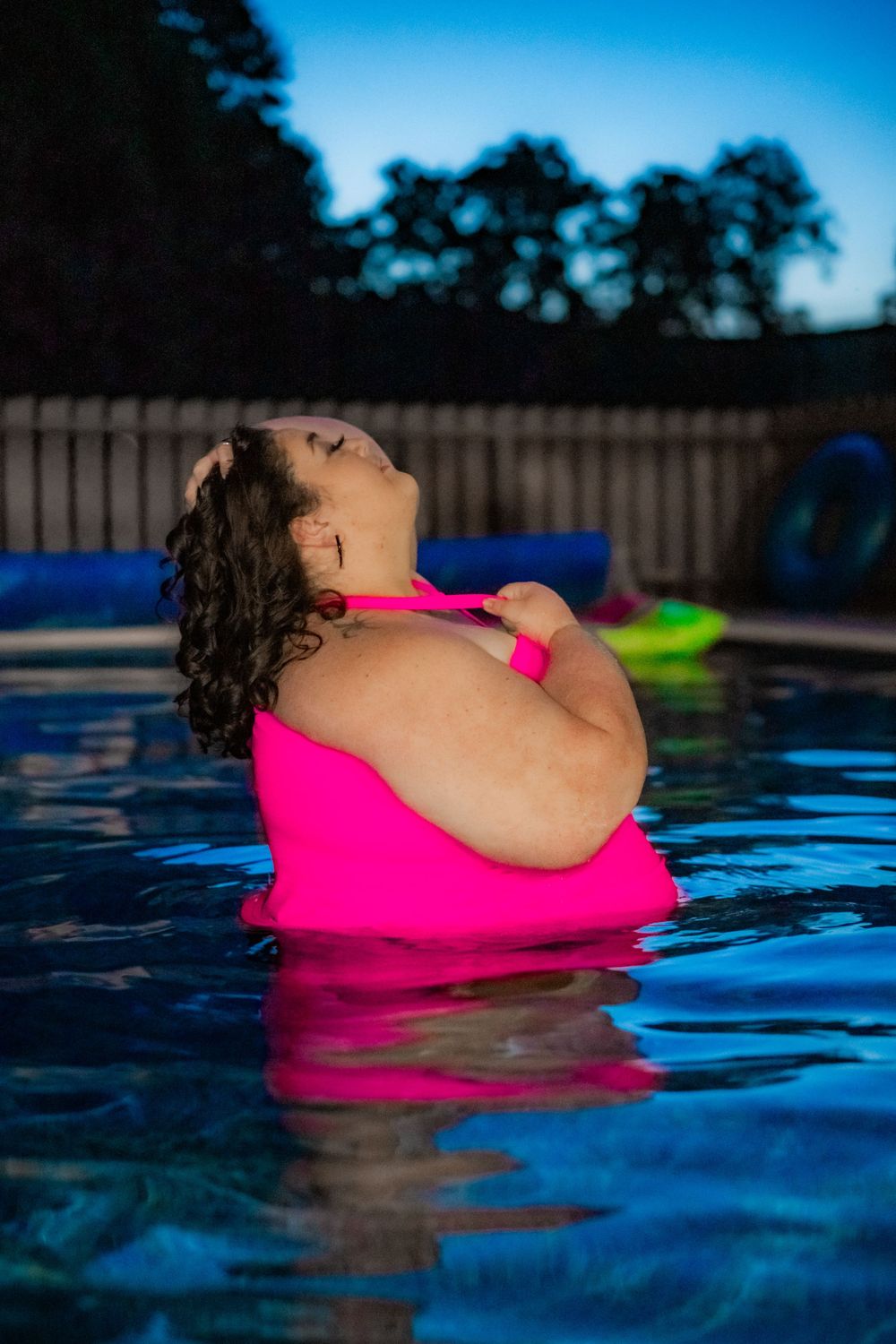 A plus-size woman in a pink swimsuit relaxes in a backyard pool at dusk, head tilted back peacefully. Pool Boudoir. Poolside Boudoir.