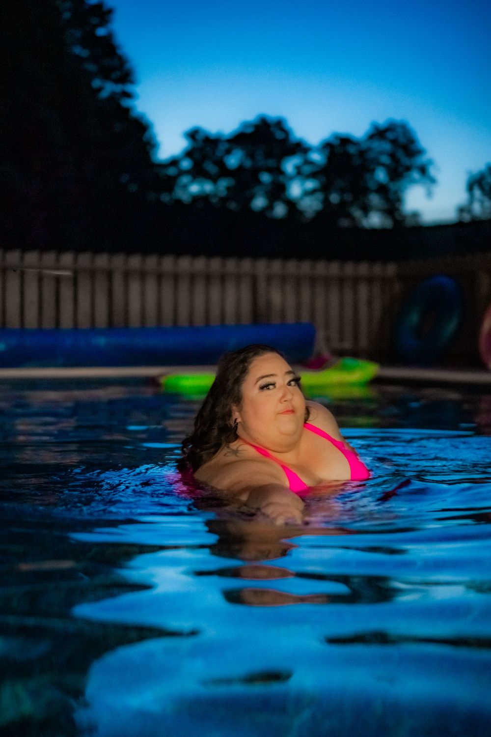 Woman in pink bikini relaxing in illuminated pool at dusk with colorful floats and trees in background. Pool Boudoir. Poolside Boudoir.