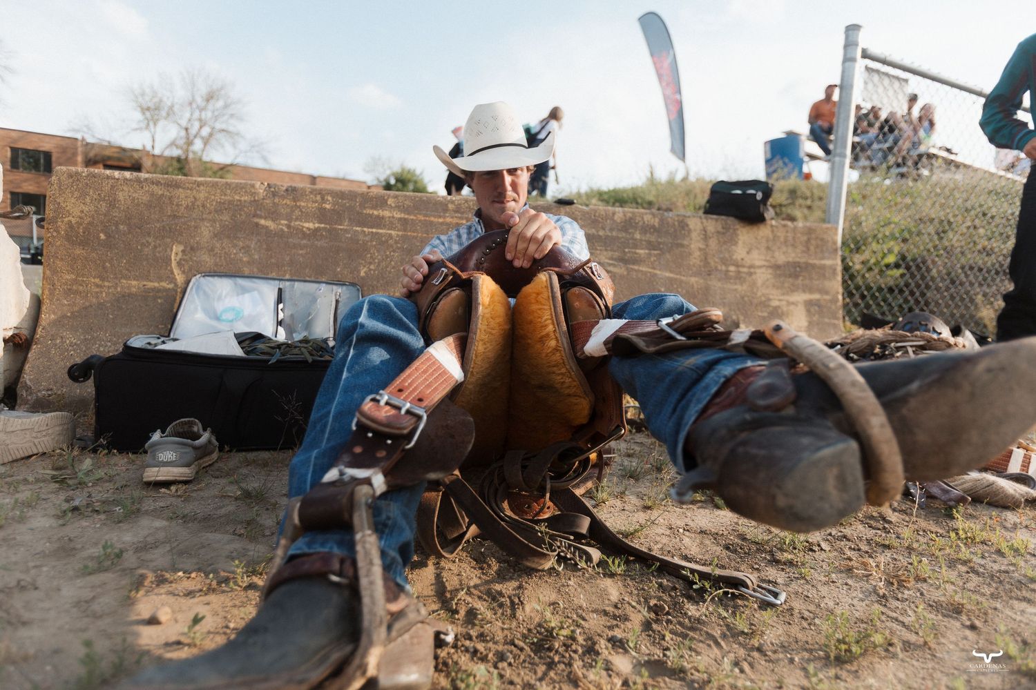 Cowboy in white helmet sits on ground at rodeo event, wearing leather chaps and protective gear outdoors.
