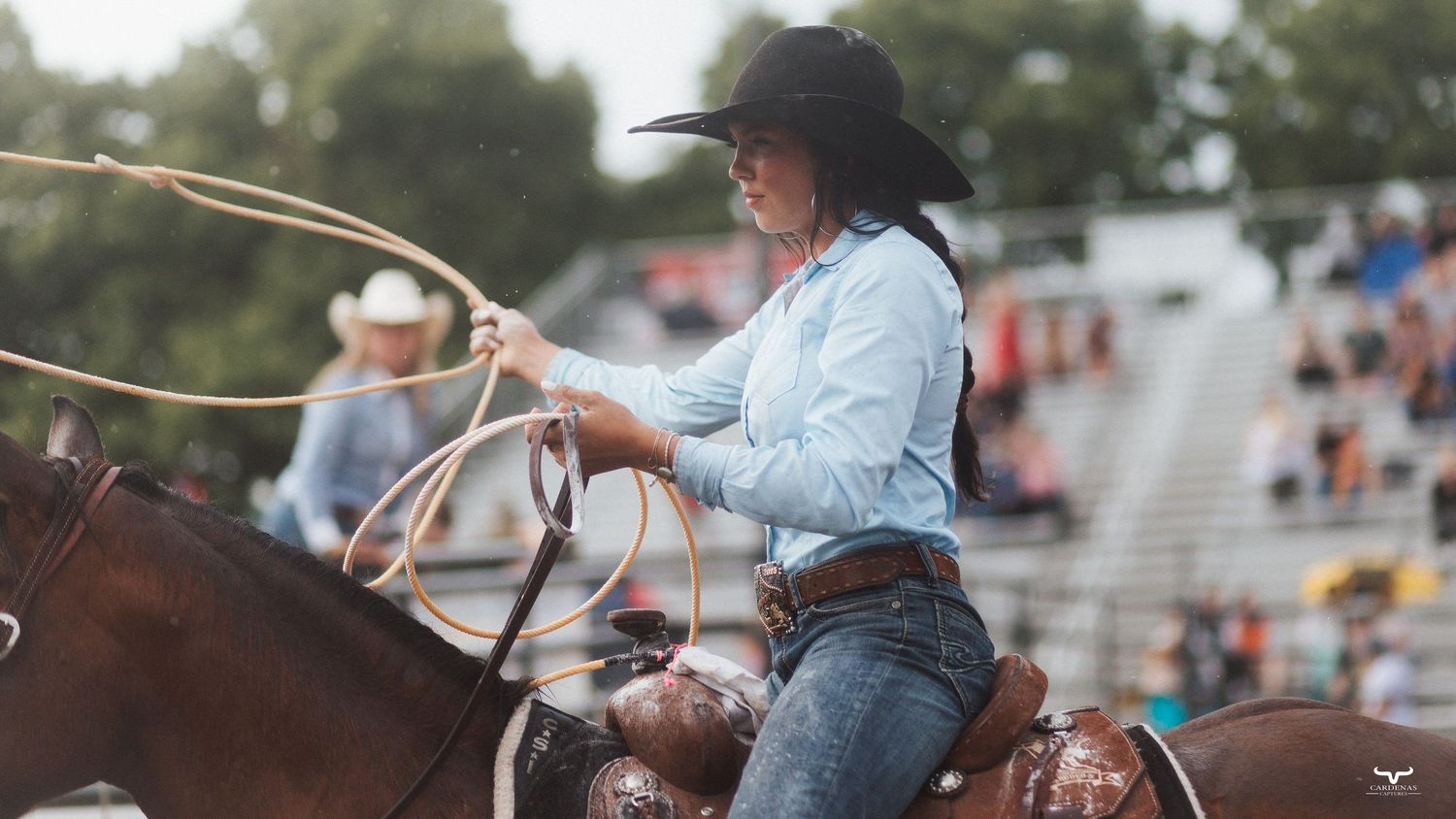 A cowgirl on horseback swings a lasso at an outdoor rodeo event, wearing a black hat and blue shirt.