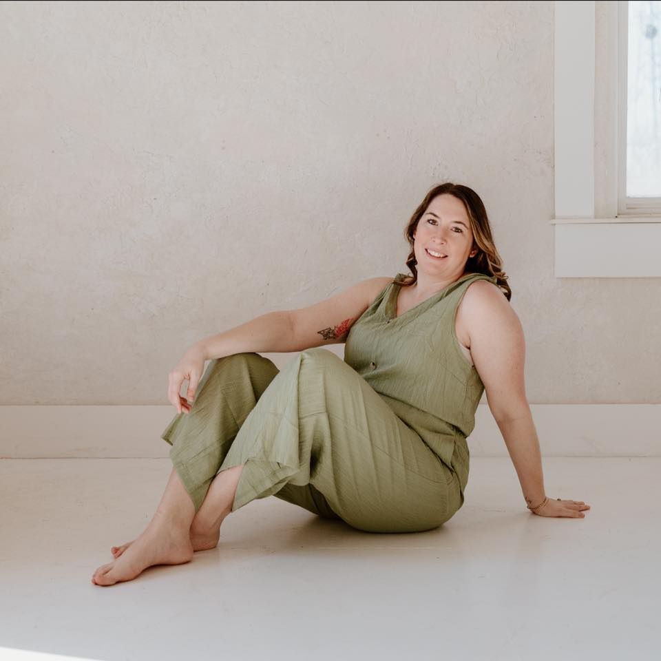 Relaxed person in olive green jumpsuit sits on white floor against neutral wall in natural lighting.