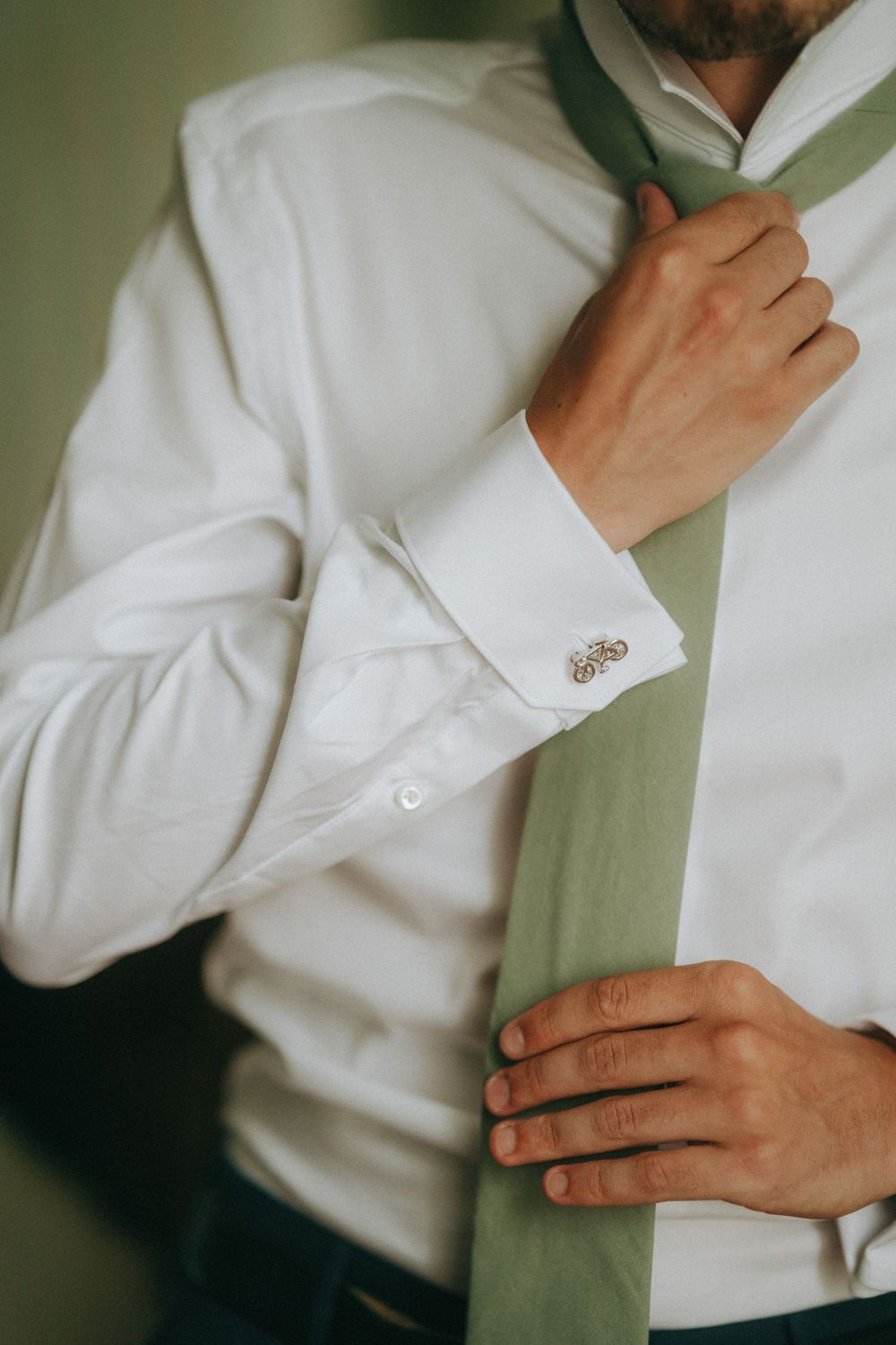 Man in white dress shirt with cufflinks adjusting a sage green tie, getting dressed for a formal occasion.