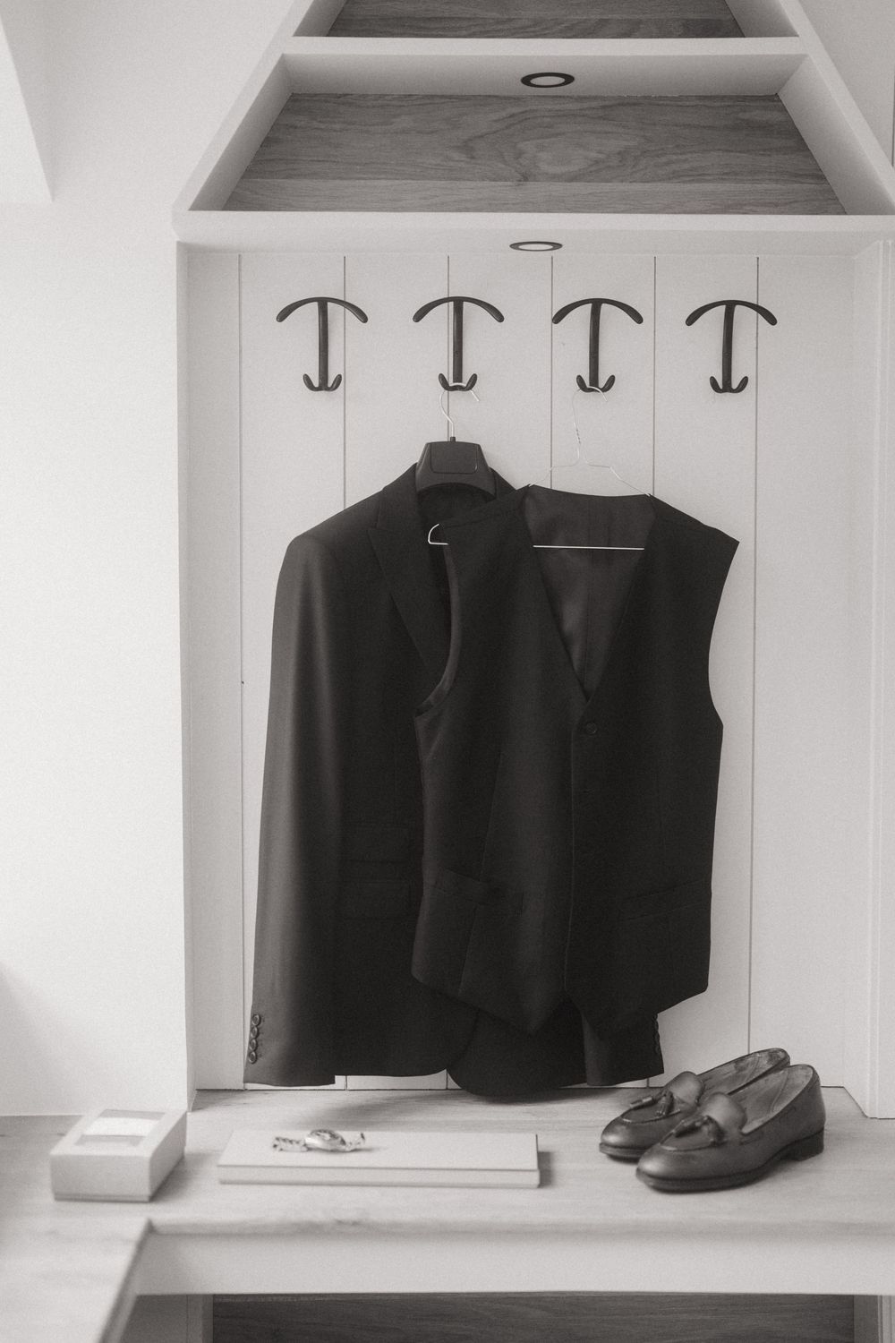 Black suit jacket and vest hanging on wall hooks beside leather loafers and accessories in monochrome.