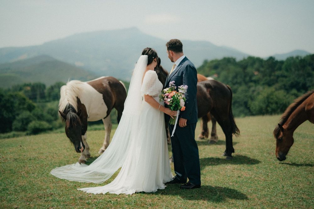 Bride and groom share a romantic moment in a green field with grazing horses and mountain backdrop.
