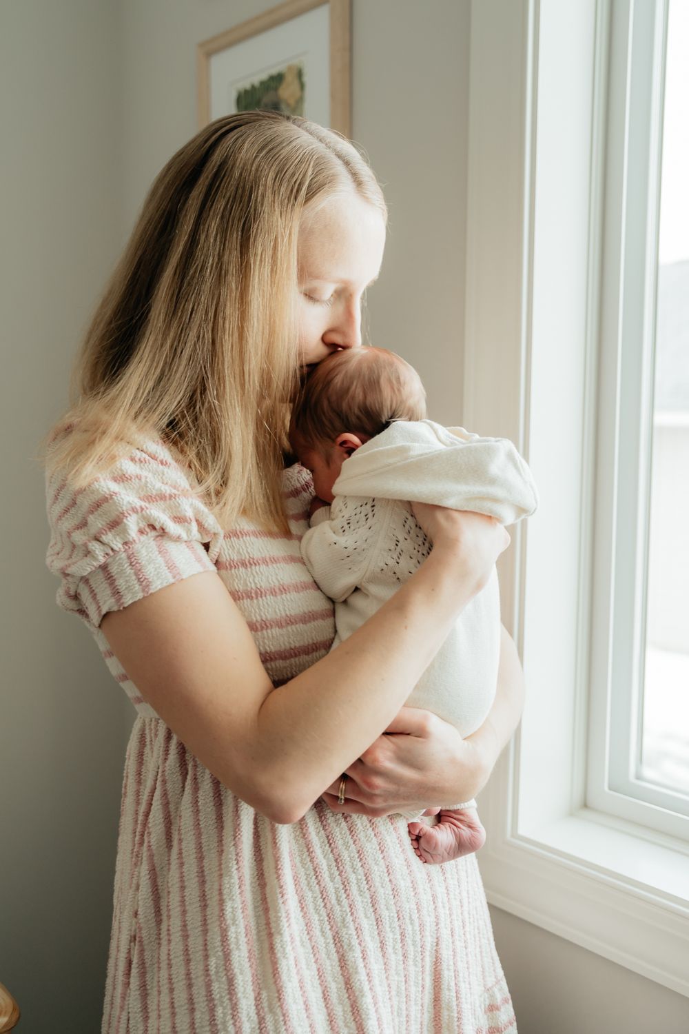 A tender moment between parent and newborn by a sunlit window in a striped dress.