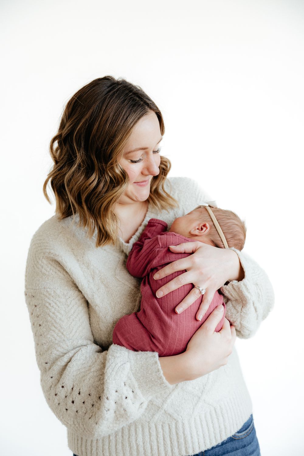 A woman in a cream sweater lovingly gazes at her newborn baby wearing burgundy against a white backdrop.