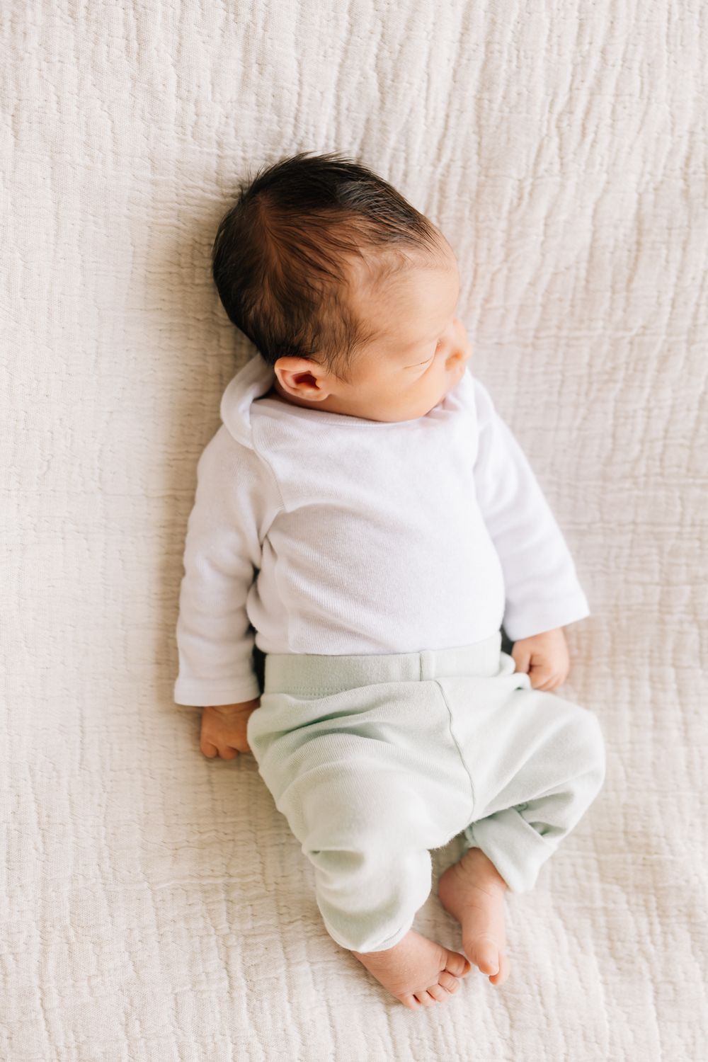 A sleeping baby in white clothing lies on a textured white blanket.