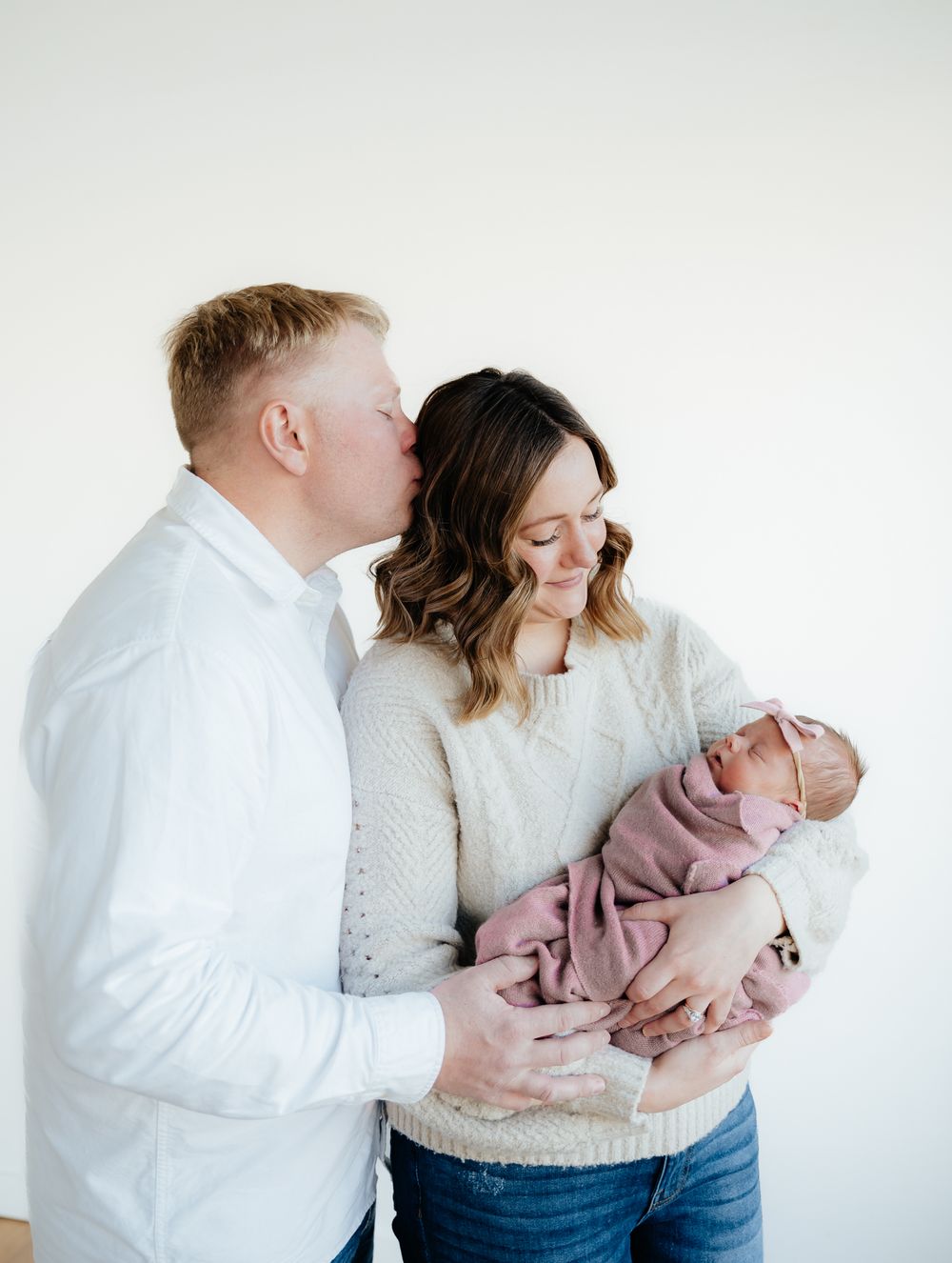 A tender family moment as parents in white and cream sweaters hold their newborn baby against a light background.