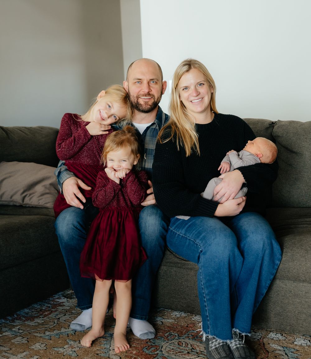 Family sits together on a grey couch, sharing casual moments in denim and burgundy clothing.