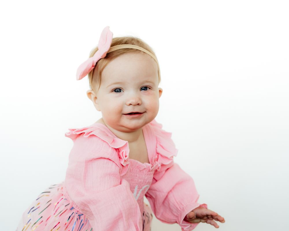 Baby wearing pink ruffled dress and headband smiles while crawling on white background.