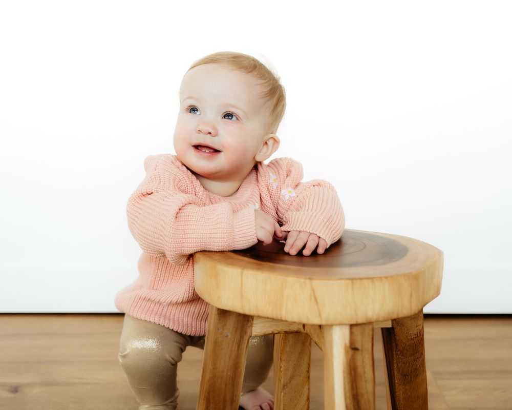 A baby in a peach-colored sweater leans on a wooden stool while smiling against a white background.