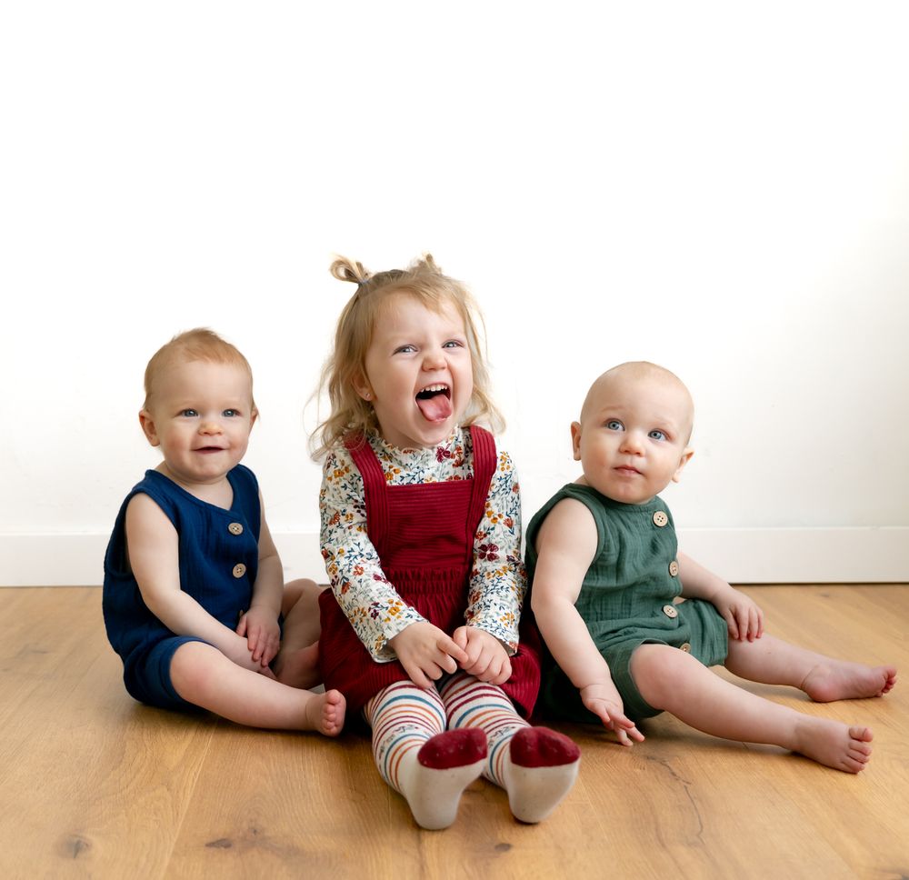 Three babies sitting together on a wooden floor wearing coordinated casual outfits.