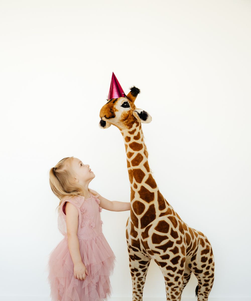 Young child in pink tulle dress standing next to tall decorative giraffe wearing party hat.