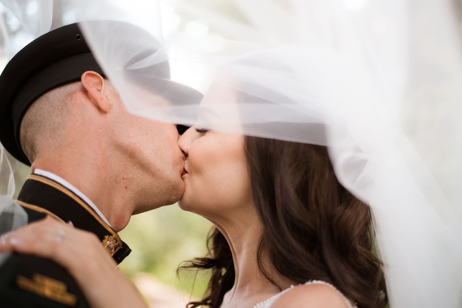 Military couple sharing romantic kiss under bridal veil at outdoor wedding.