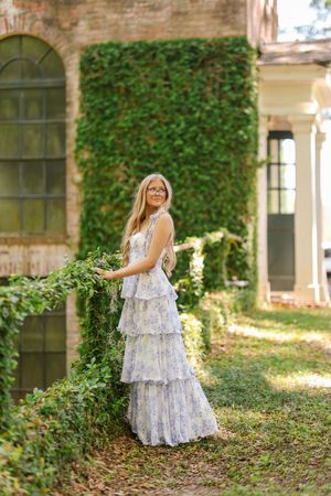 A tiered white lace dress stands elegantly in a garden setting with ivy-covered walls and dappled sunlight.