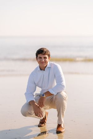 A person wearing light colored clothing crouching on a sandy beach at sunset.