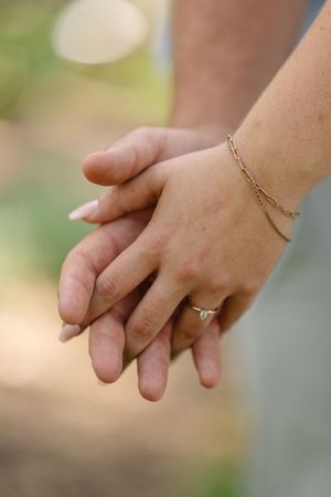 Closeup of hands with delicate gold bracelet and engagement ring against blurred outdoor background.